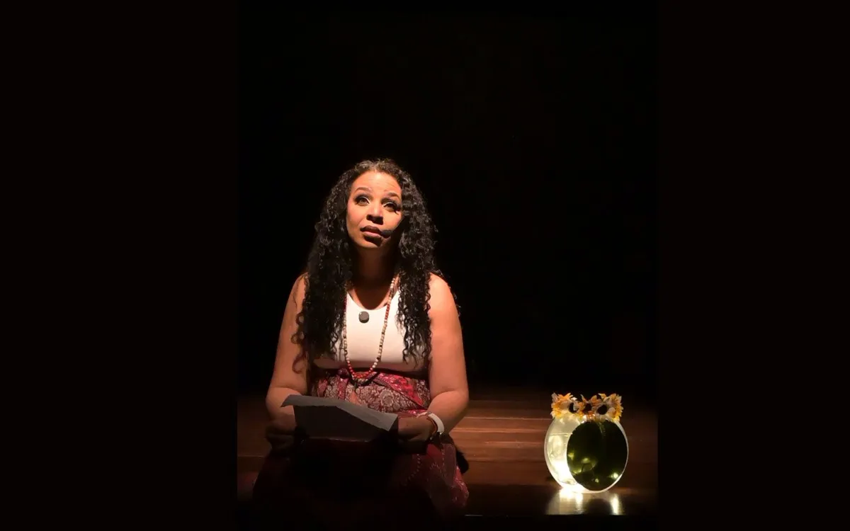 Mujer sentada en el escenario bajo una luz cálida, sosteniendo papeles en sus manos, con expresión reflexiva. A su lado, un florero iluminado con girasoles decora la escena. El fondo es oscuro, destacando la atmósfera íntima y emocional del momento.